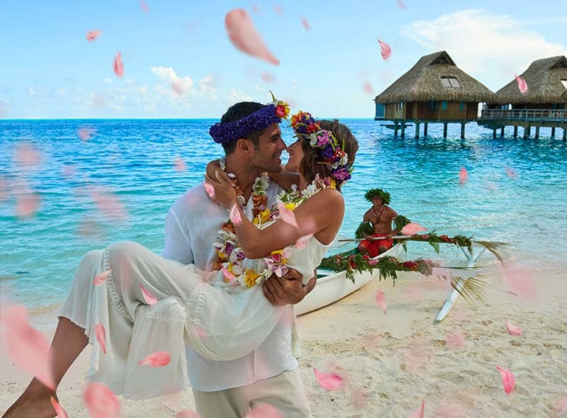 Et par som gifter seg på stranda på Bora Bora med vannvillaer i bakgrunnen.
