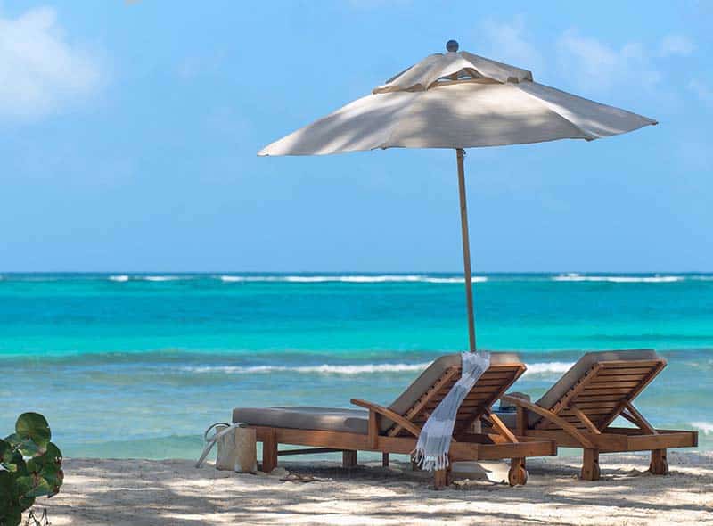 To solstoler i tre med tykke puter og en hvit parasol på en sandstrand med turkist hav i bakgrunnen.